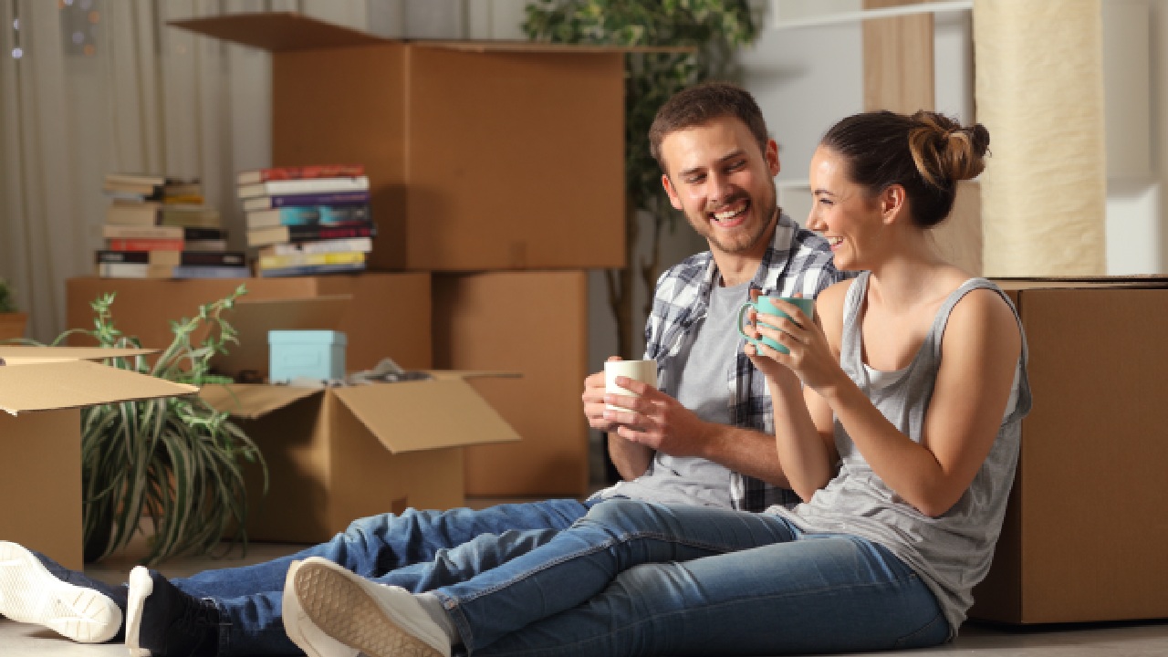 A happy Australian couple taking a break while moving house, sitting on the floor surrounded by boxes, coffee cups in hand. Perfect depiction of moving house interstate in Australia.