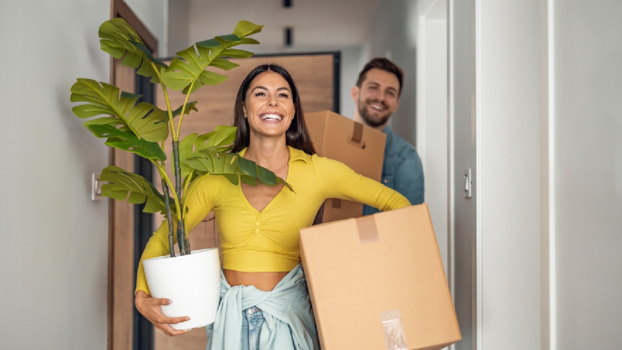 Smiling couple carrying moving boxes and a houseplant into a new home, demonstrating packing the house to move in an organised and positive way